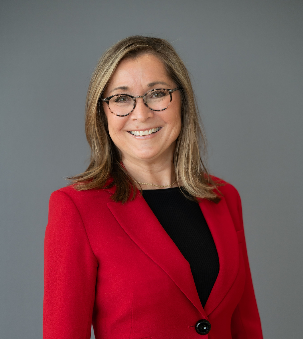 Portrait of Leslie J Willians, A member of Axely'fs Board of Directors; a woman with light brown hair, wearing glasses, a red blazer, and a black top, smiling against a gray background.