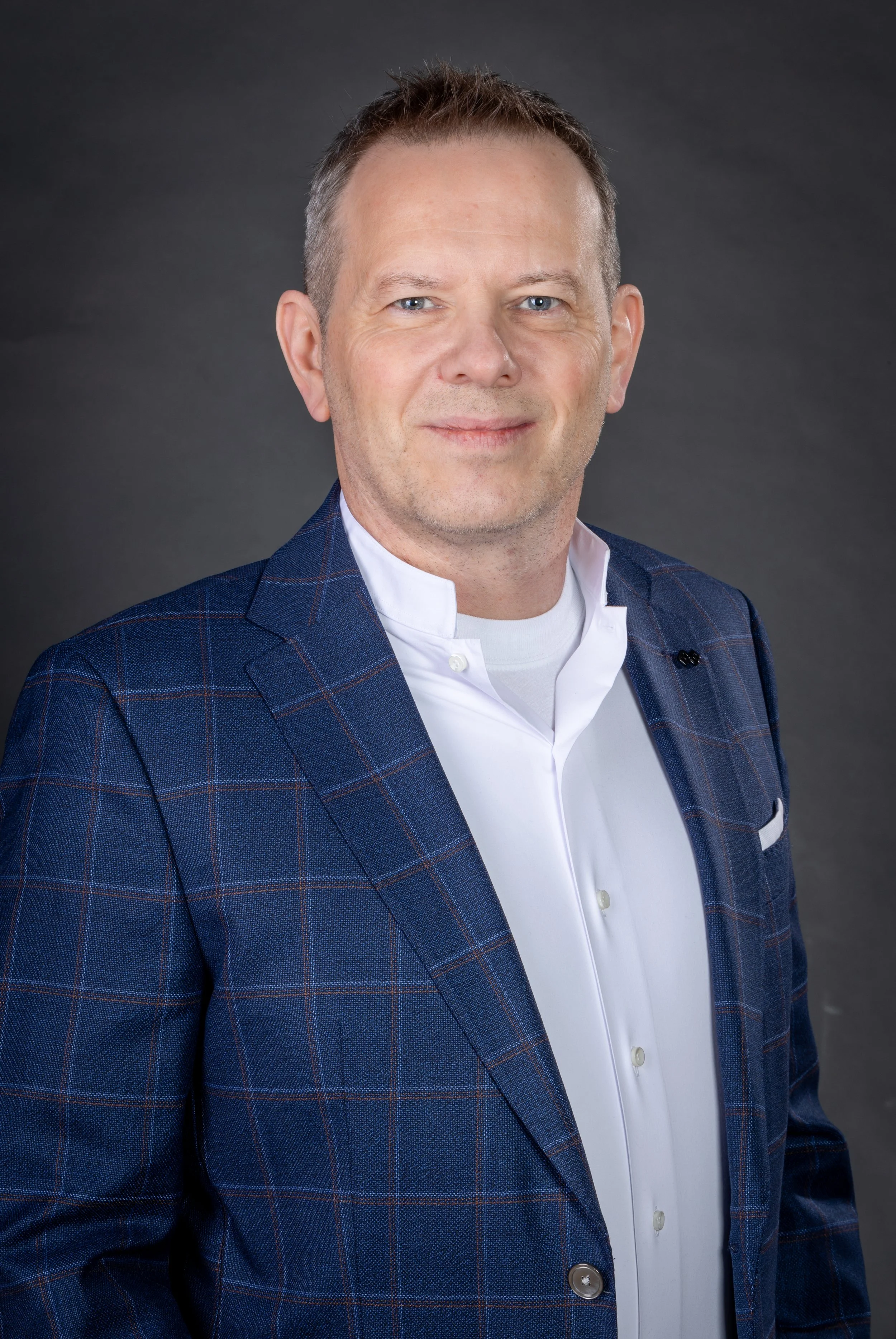 A man in a blue checkered blazer and white shirt posing against a dark grey background
