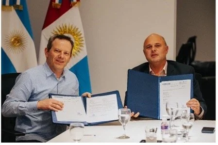 Two men sitting at a table, holding open documents, with flags of Argentina and another country in the background.