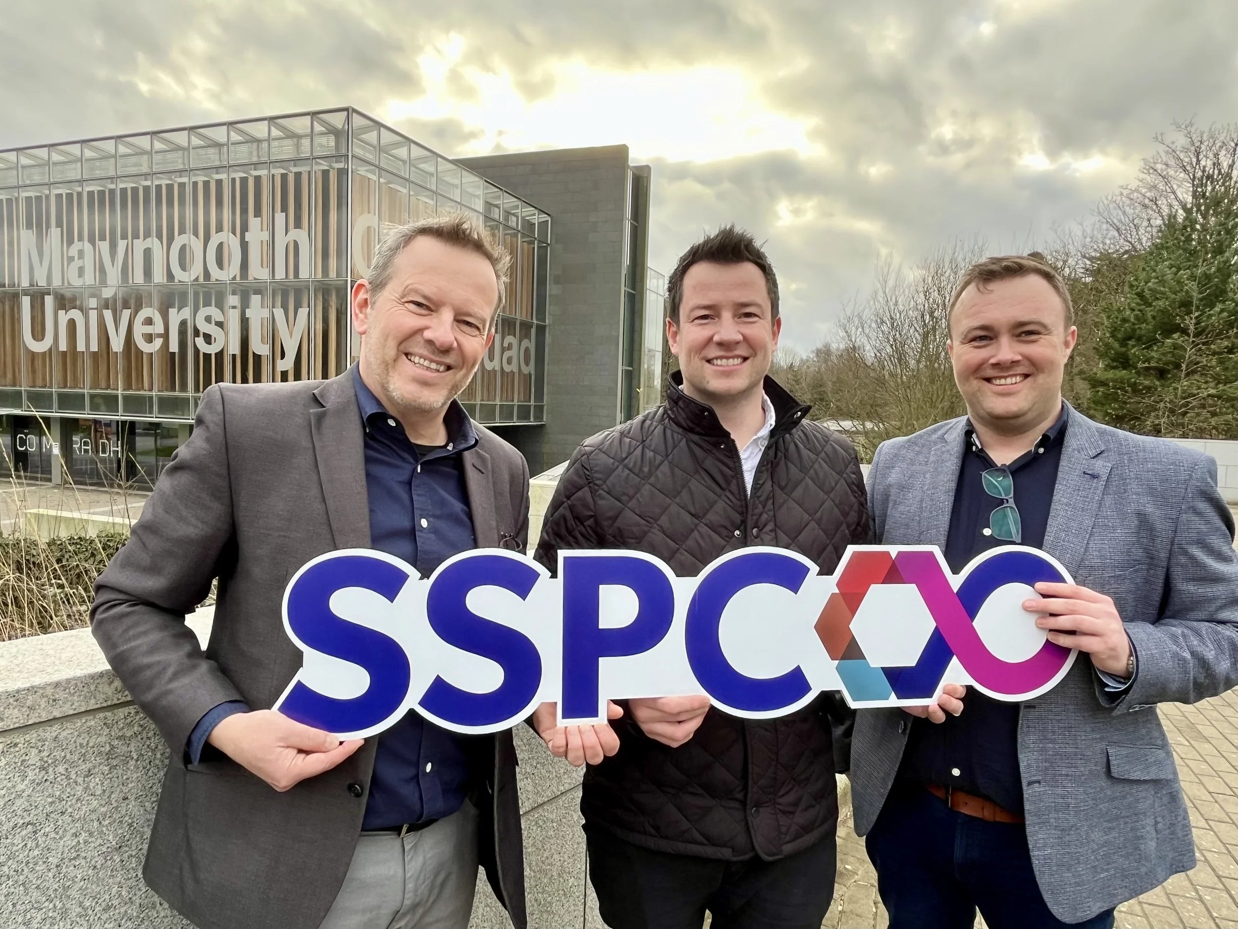 Three men in business attire holding a sign with the acronym SSPCX in front of Maynooth University campus during daytime with cloudy sky.